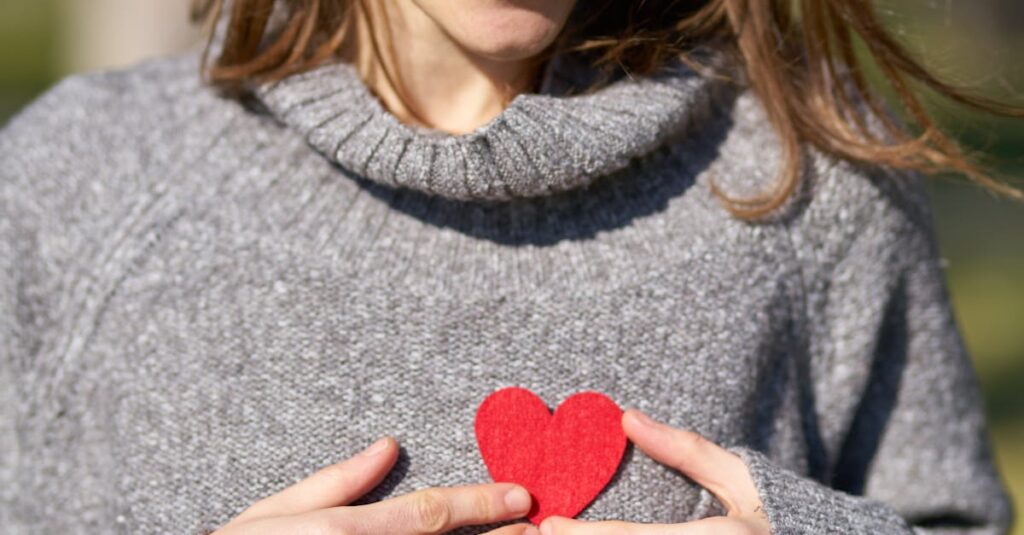 A woman outdoors holding a red heart against her chest, capturing a warm moment.