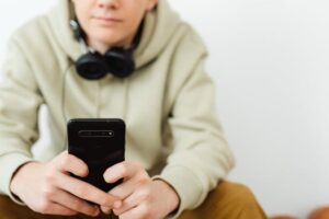 Teenager in hoodie uses smartphone and headphones indoors.