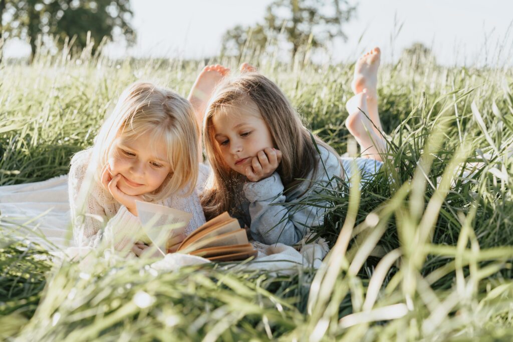 Two young girls lying in a grassy field reading a book together on a sunny day.