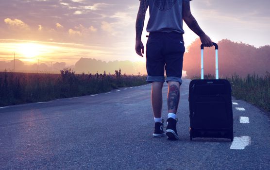 Man with tattoo walks along a rural road at sunrise with a suitcase, showcasing travel and adventure.