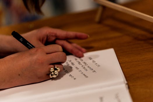 Handwriting in a notebook on a wooden table with a focus on a pen and ringed hand.