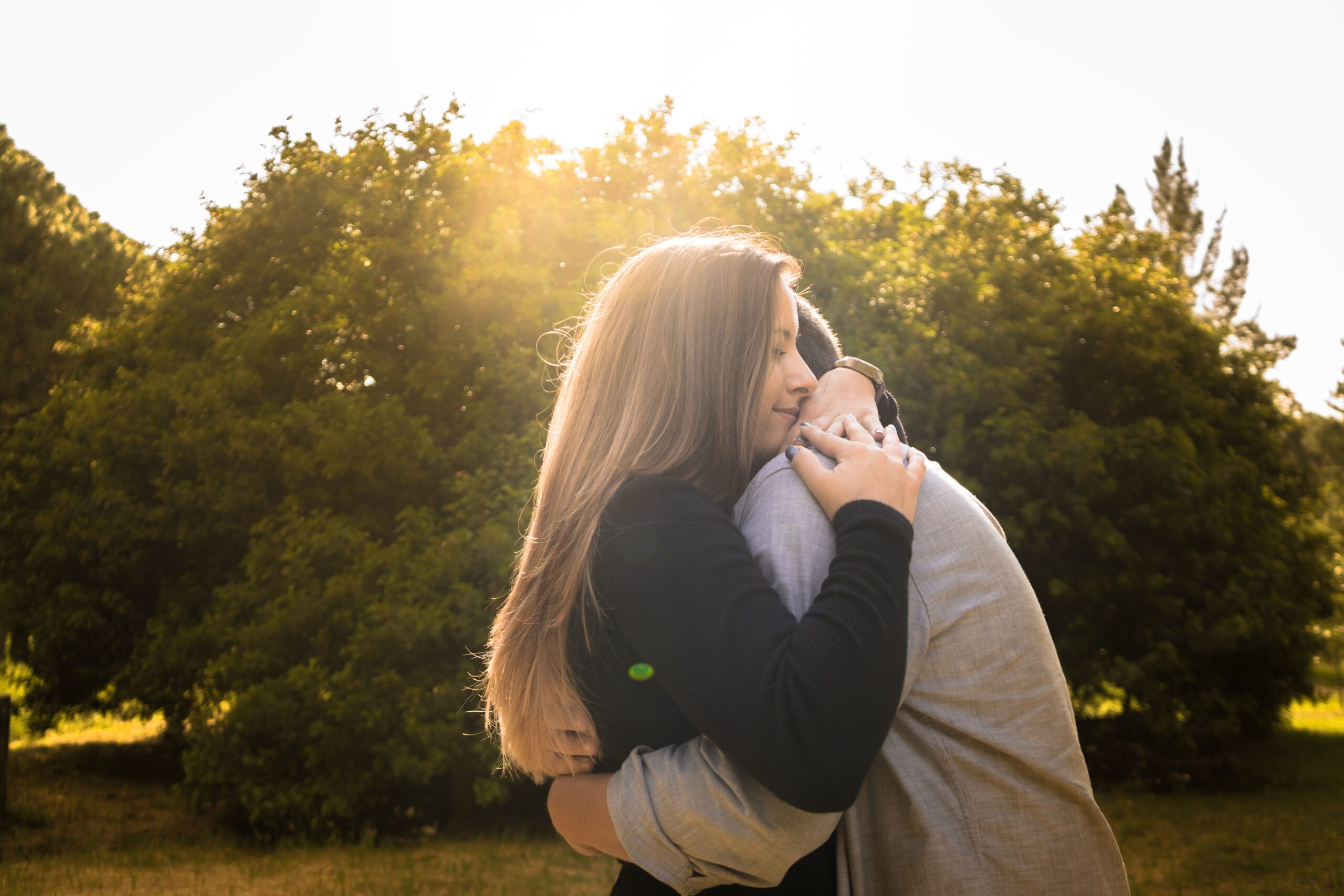 A couple shares a warm embrace under the bright sun in a peaceful park setting.