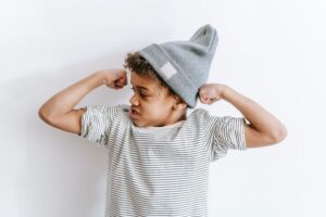 Strong African American boy in stylish hat showing biceps while standing on white background with arms bent in elbows in light room