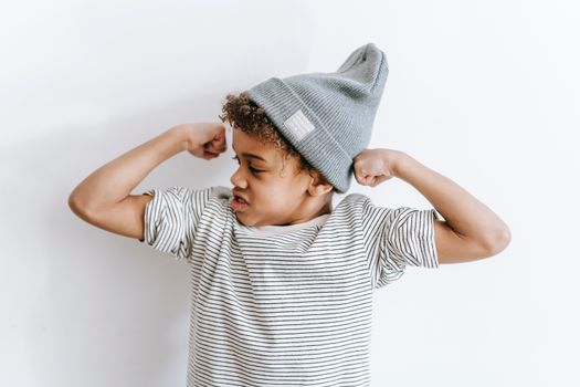 Strong African American boy in stylish hat showing biceps while standing on white background with arms bent in elbows in light room